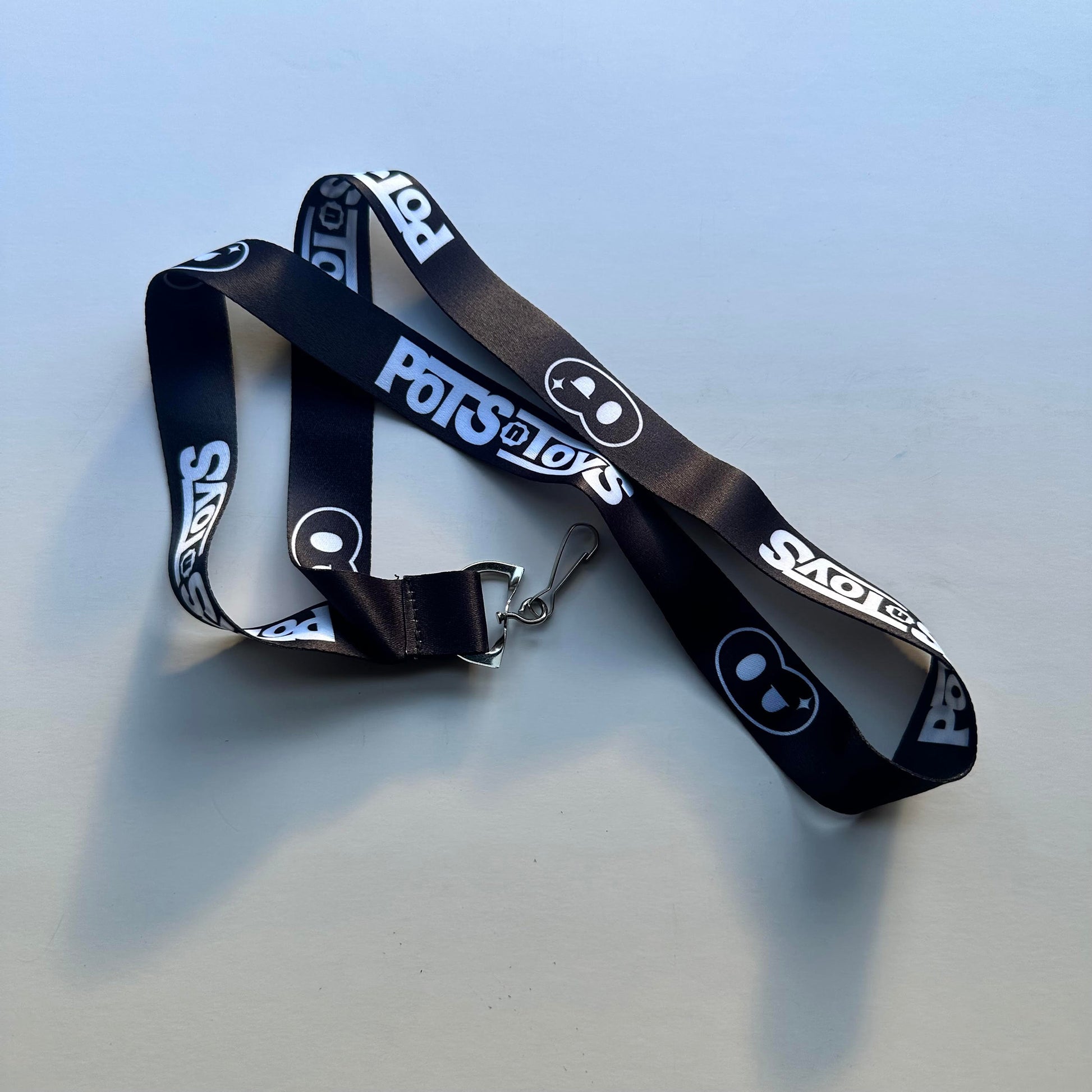 A sleek black lanyard with the “Pots@Toys” logo printed repeatedly in white, featuring a playful pot-shaped icon. The design is minimalistic and professional, making it ideal for conventions or promotional events.