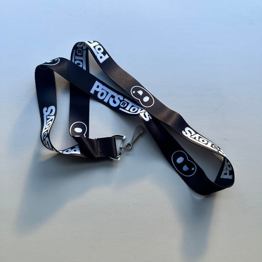 A sleek black lanyard with the “Pots@Toys” logo printed repeatedly in white, featuring a playful pot-shaped icon. The design is minimalistic and professional, making it ideal for conventions or promotional events.